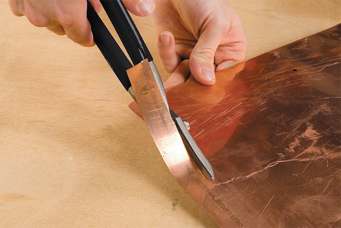 Eine Person schneidet mit einer Metallschere ein dünnes Kupferblech auf einem Holztisch.