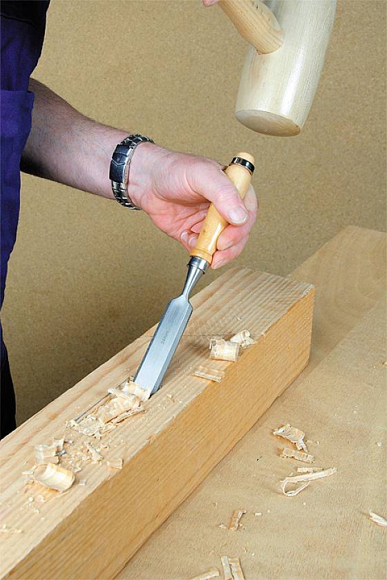 Eine Person schnitzt mit einem Stechbeitel und einem Holzhammer Holzspäne von einem Holzblock ab.