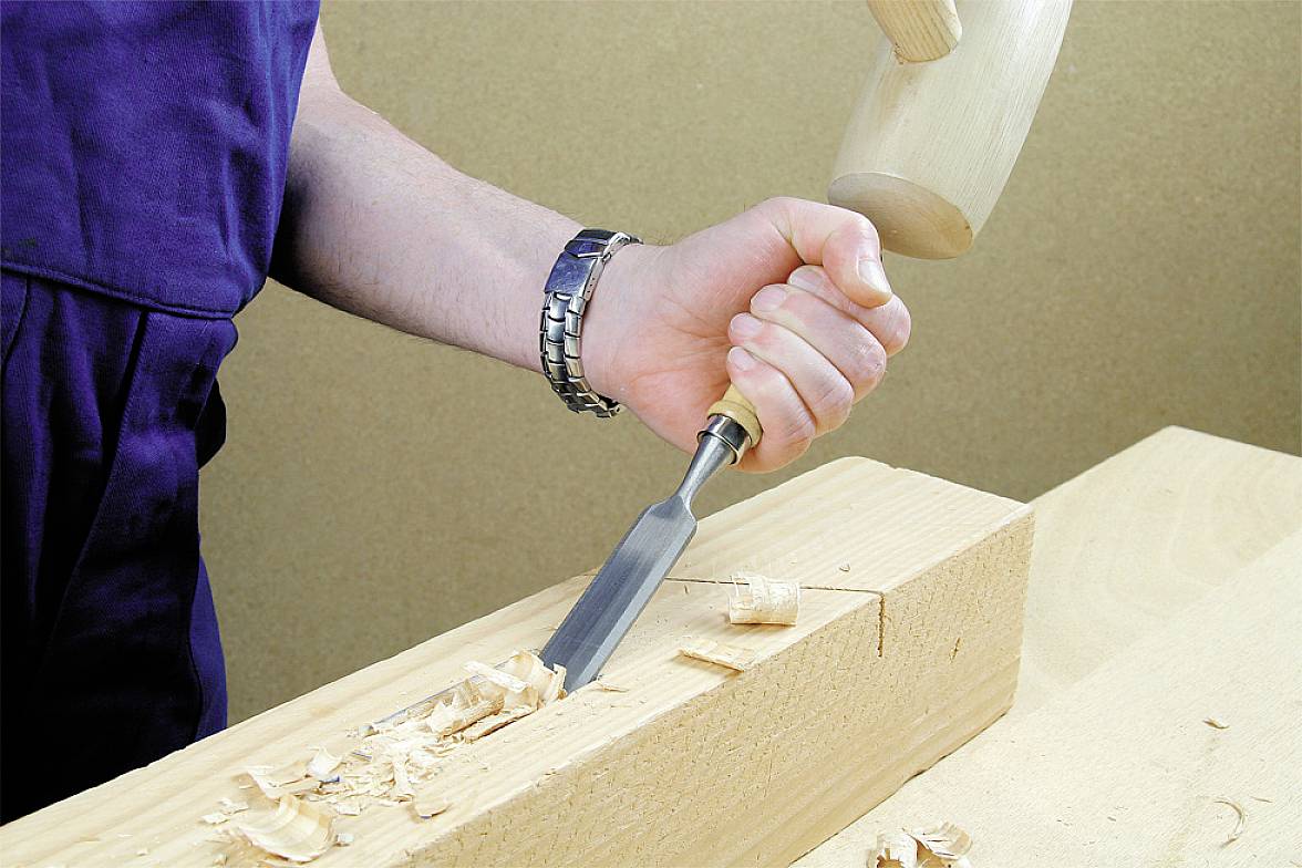 Eine Person schnitzt mit einem Meißel und Hammer Holzspäne aus einem Holzstück. Sie trägt ein blaues Arbeitsoutfit.
