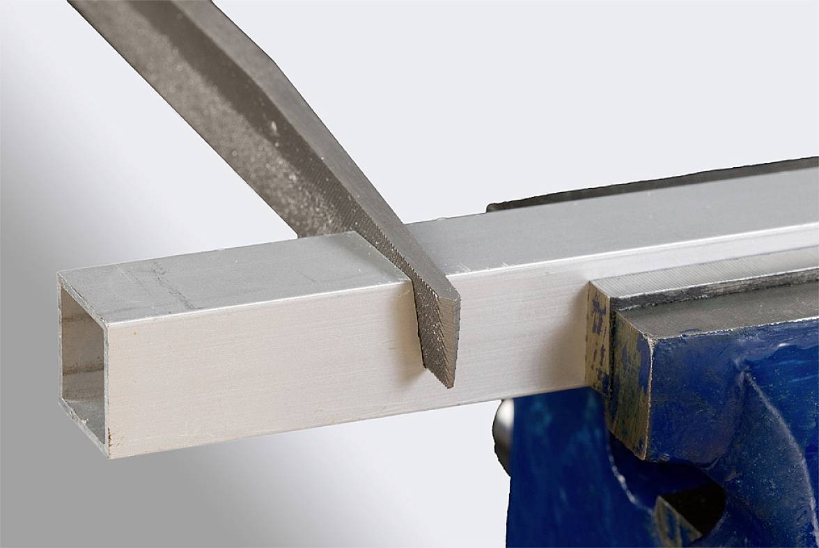 Metallfeile bearbeitet ein rechteckiges Aluminiumprofil, das in einem Schraubstock eingespannt ist. Fokus auf Präzision der Arbeit.