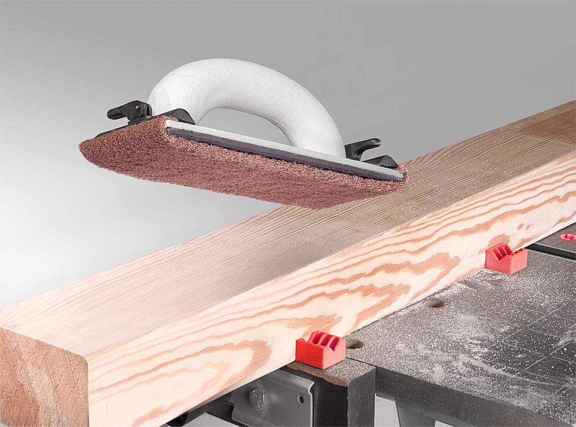 Ein Handschleifgerät bearbeitet die Oberfläche eines Holzbalkens. Der Balken liegt auf einer Werkbank, umgeben von Holzspänen.
