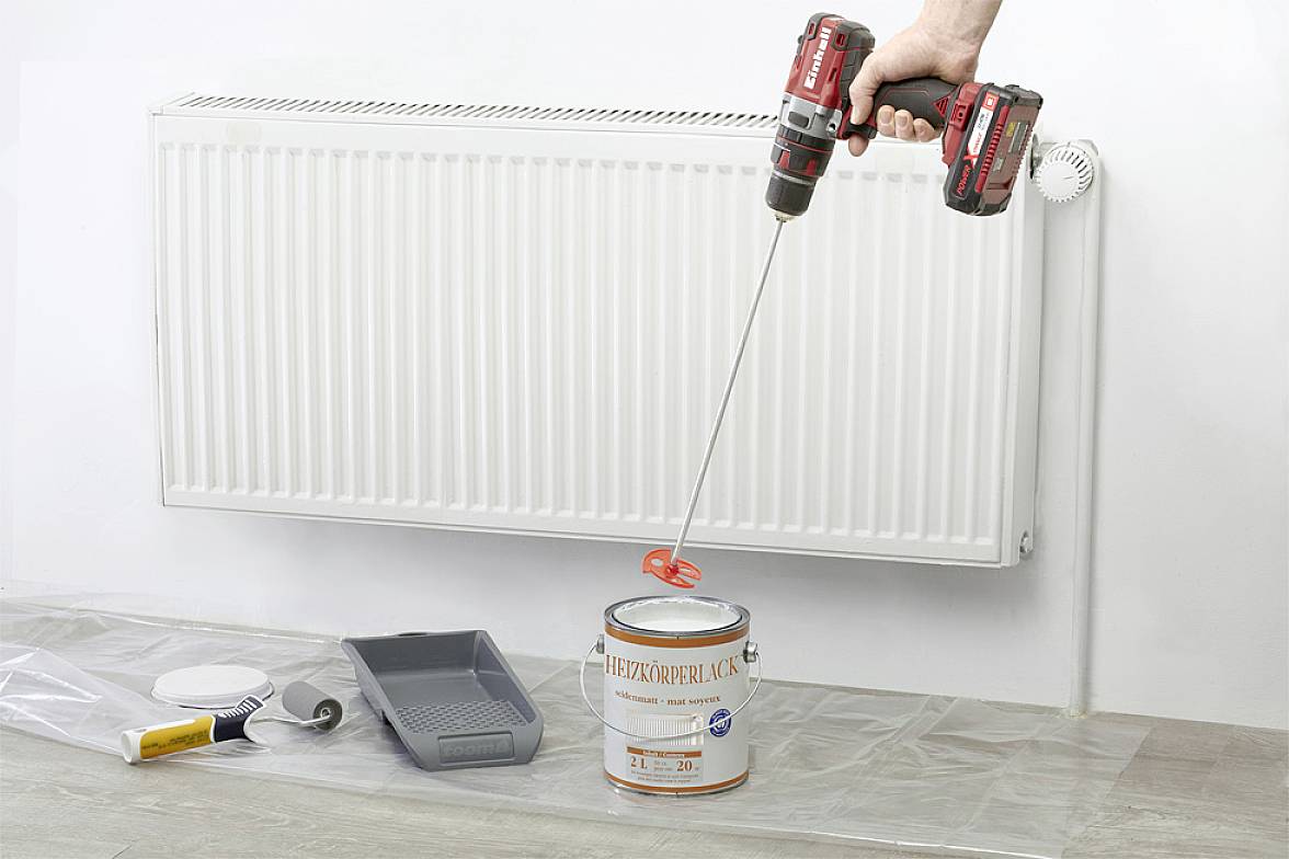 Ein Heizkörper vor einer weißen Wand. Eine Person hält einen elektrischen Mixer, der Farbe in einer offenen Dose mischt. Auf dem Boden liegen Malzubehör und -werkzeuge.