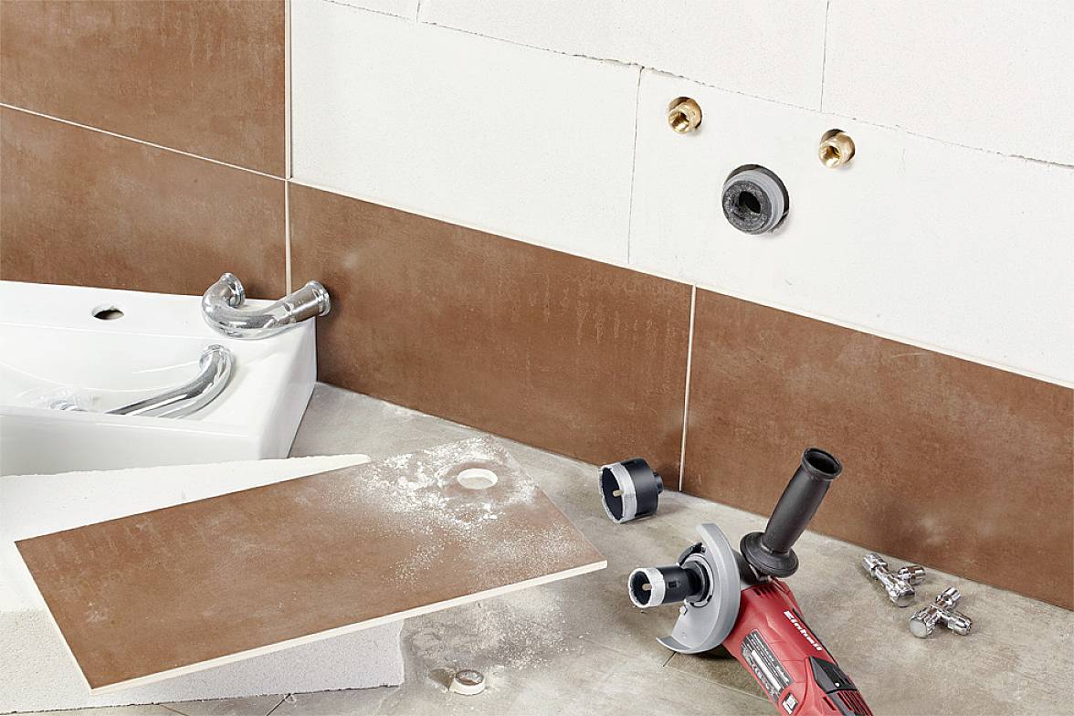 Eine Baustelle im Badezimmer zeigt halb verlegte braune Wandfliesen und ein Waschbecken. Eine rote Schleifmaschine liegt auf dem Boden.