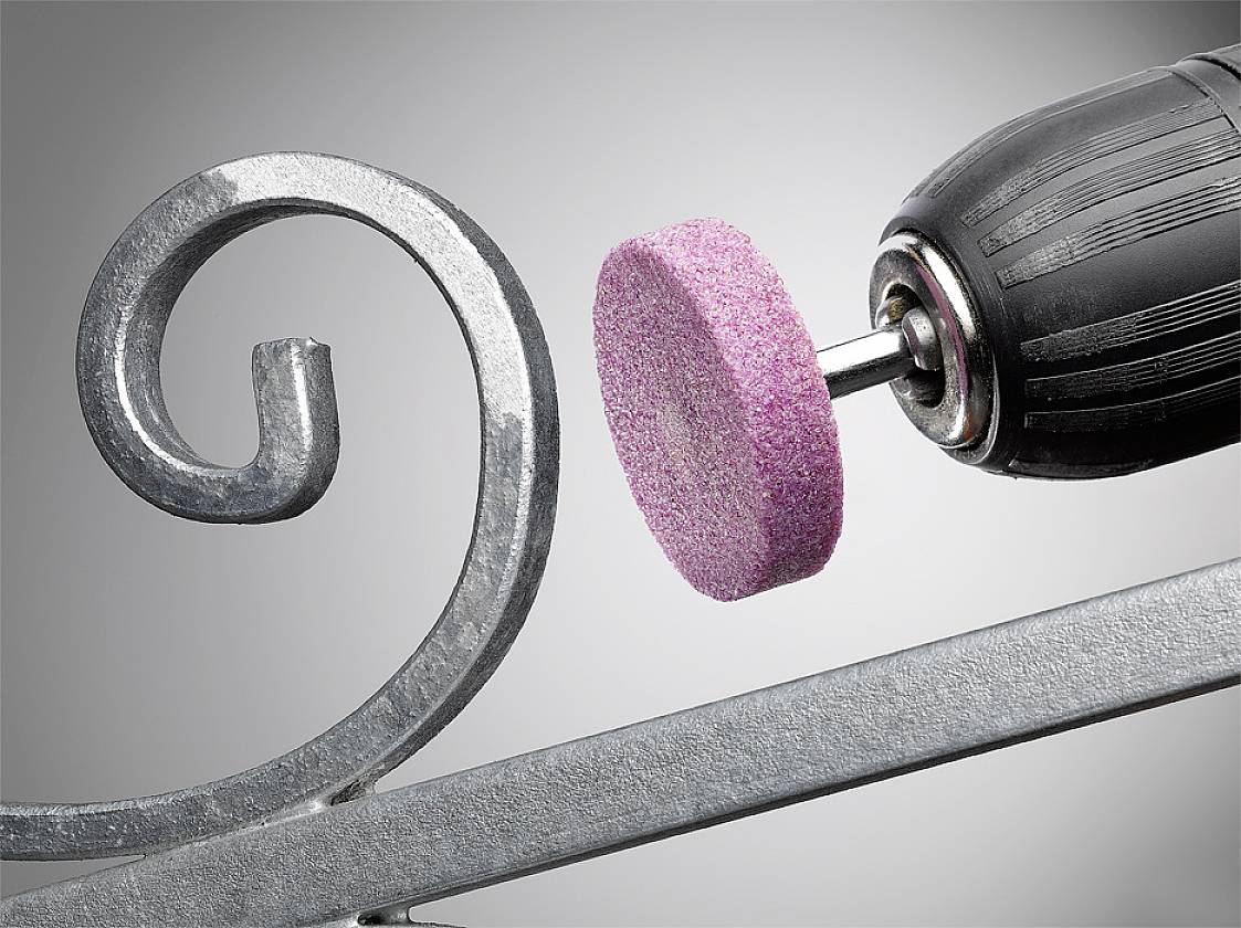Eine elektrische Bohrmaschine mit einem rosa Schleifaufsatz bearbeitet ein dekoratives Metallstück mit geschwungenem Muster.