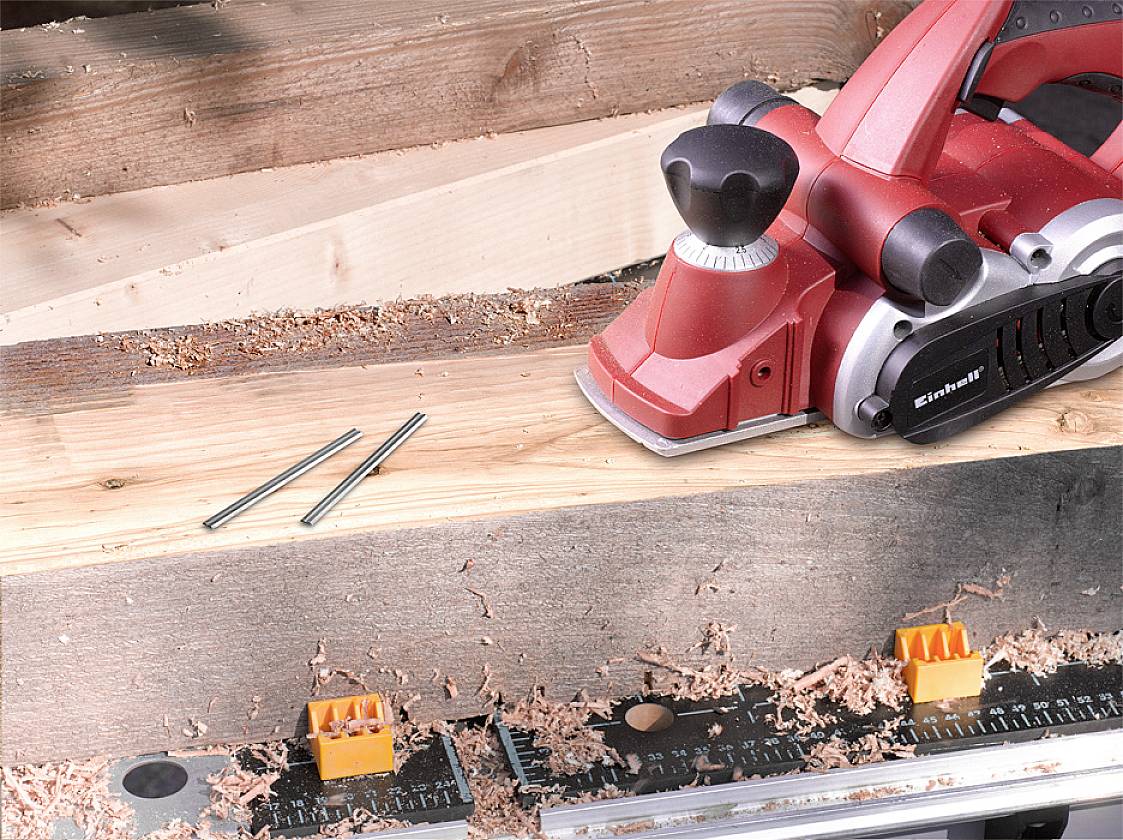 Ein roter Elektrohobel liegt auf einem Holzbrett mit Holzspänen und zwei Nägeln daneben. Eine Messleiste ist im Vordergrund sichtbar.