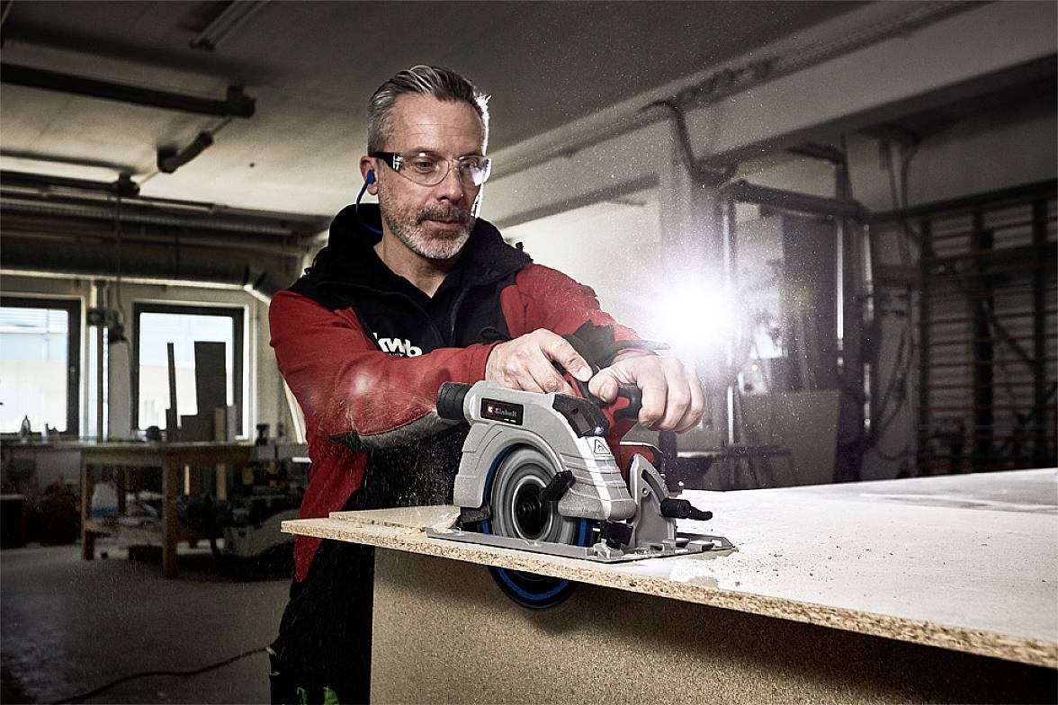 Mann mit Schutzbrille und Gehörschutz benutzt eine Handkreissäge auf einem Holztisch in einer Werkstatt.lichtdurchflutetener Raum.