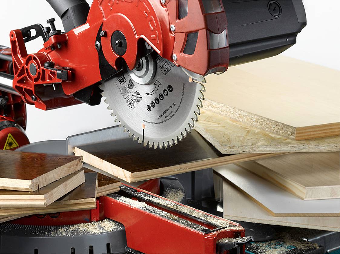 Eine elektrische Kreissäge schneidet mehrere Holzplatten unterschiedlicher Dicke und Textur. Holzspäne liegen um die Säge.