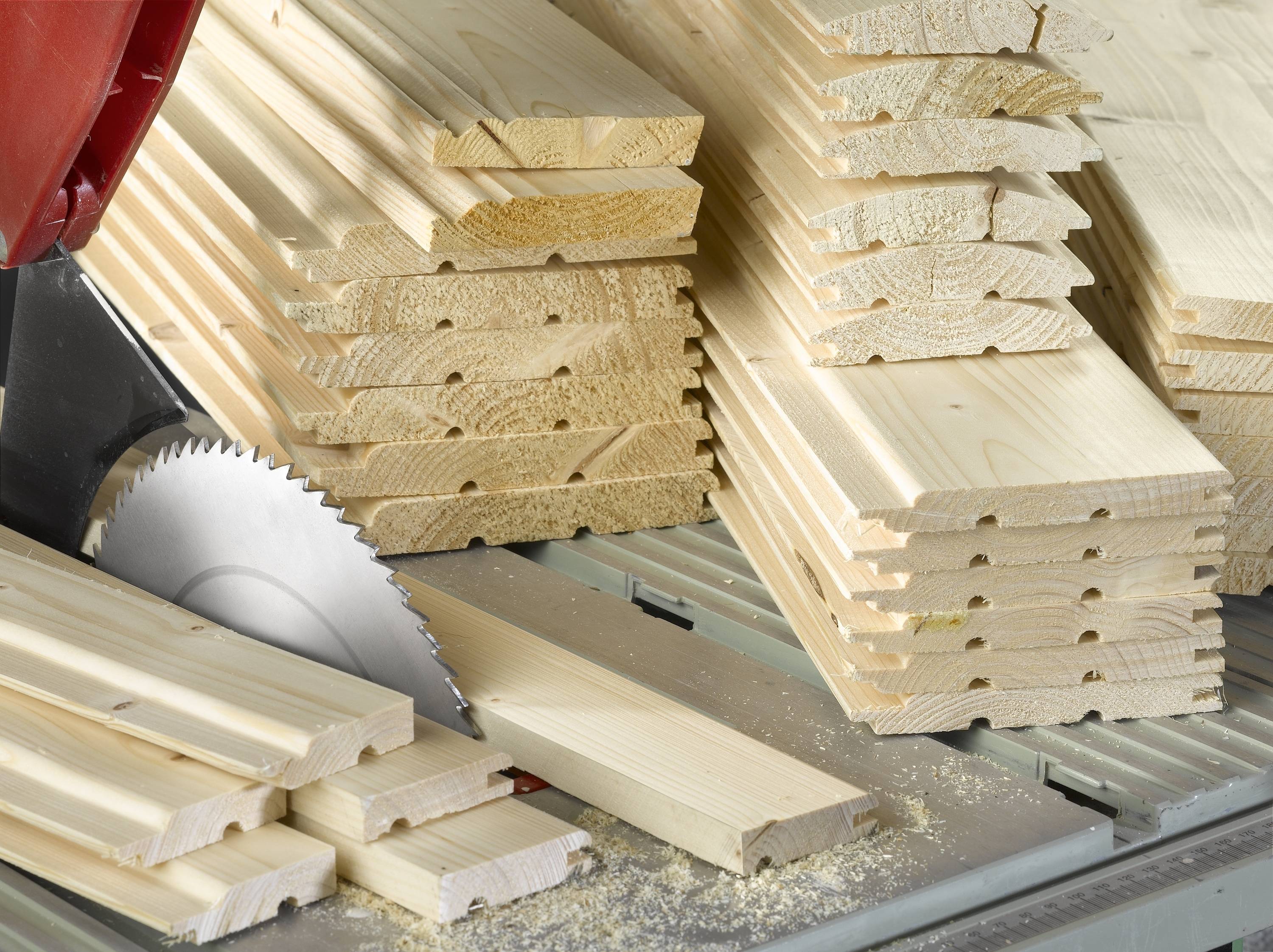 Gestapelte Holzpaneele neben einer Kreissäge in einer Werkstatt. Holzspäne auf dem Tisch deuten auf kürzliche Holzbearbeitung hin.