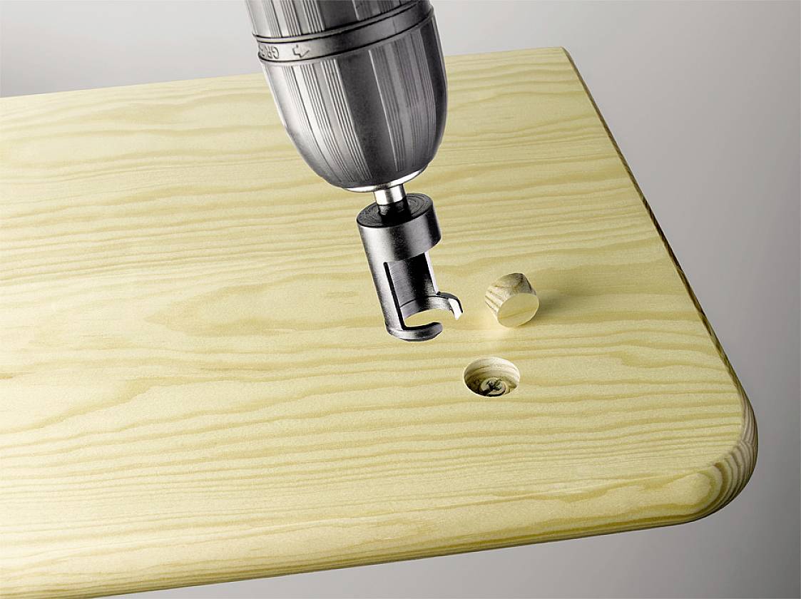 Eine Bohrmaschine mit einem Plug Cutter erzeugt zylindrische Holzstifte aus einem Holzbrett. Ein fertiger Stift liegt daneben.