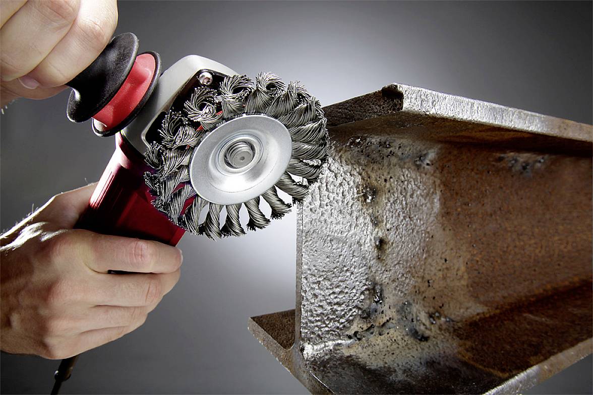 Nahaufnahme von zwei Händen, die eine elektrische Schleifmaschine mit Drahtbürste halten, um Rost von einem großen Metallträger zu entfernen.