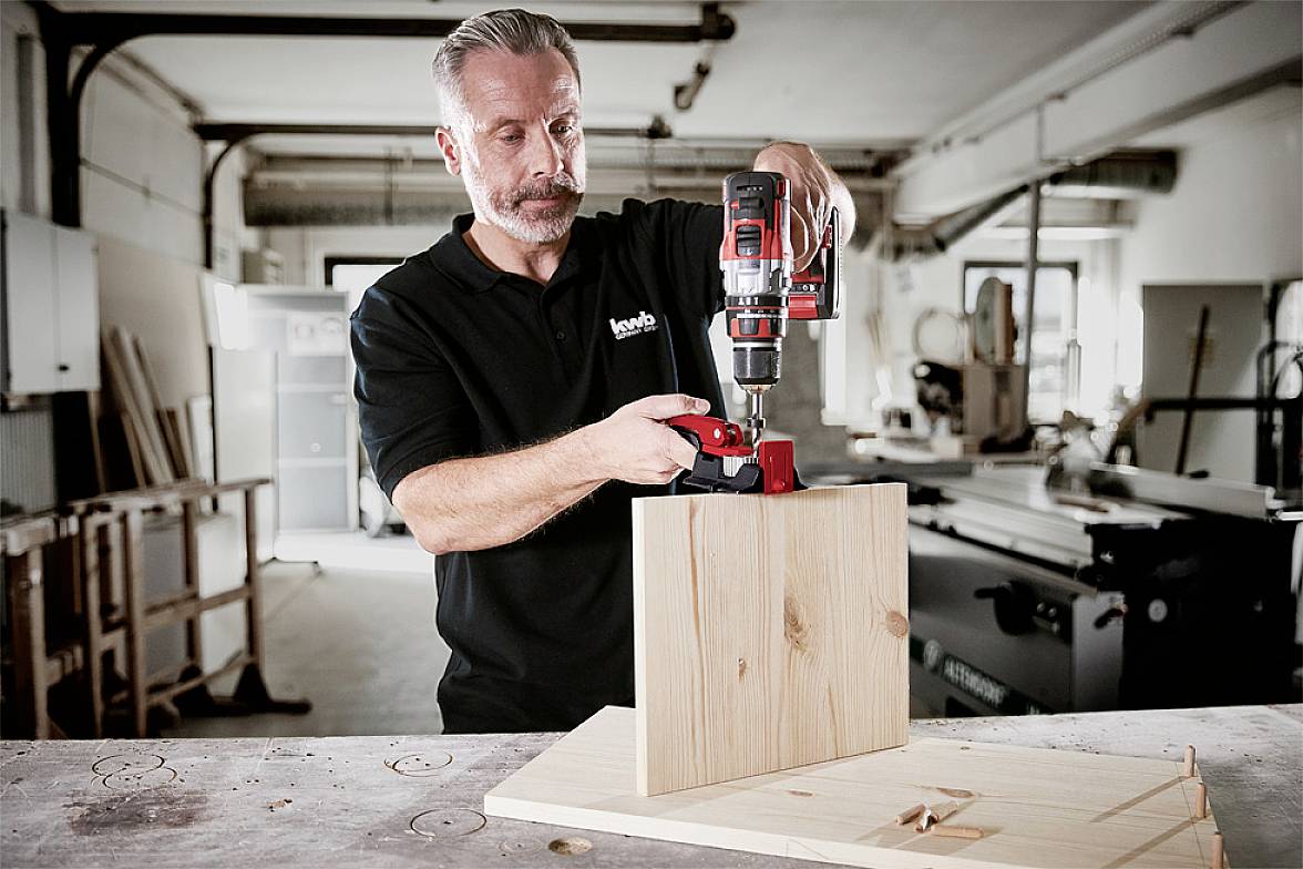 Ein Mann in einer Werkstatt benutzt einen Akkubohrer, um zwei Holzbretter zu verbinden. Im Hintergrund sind Werkzeuge zu sehen.