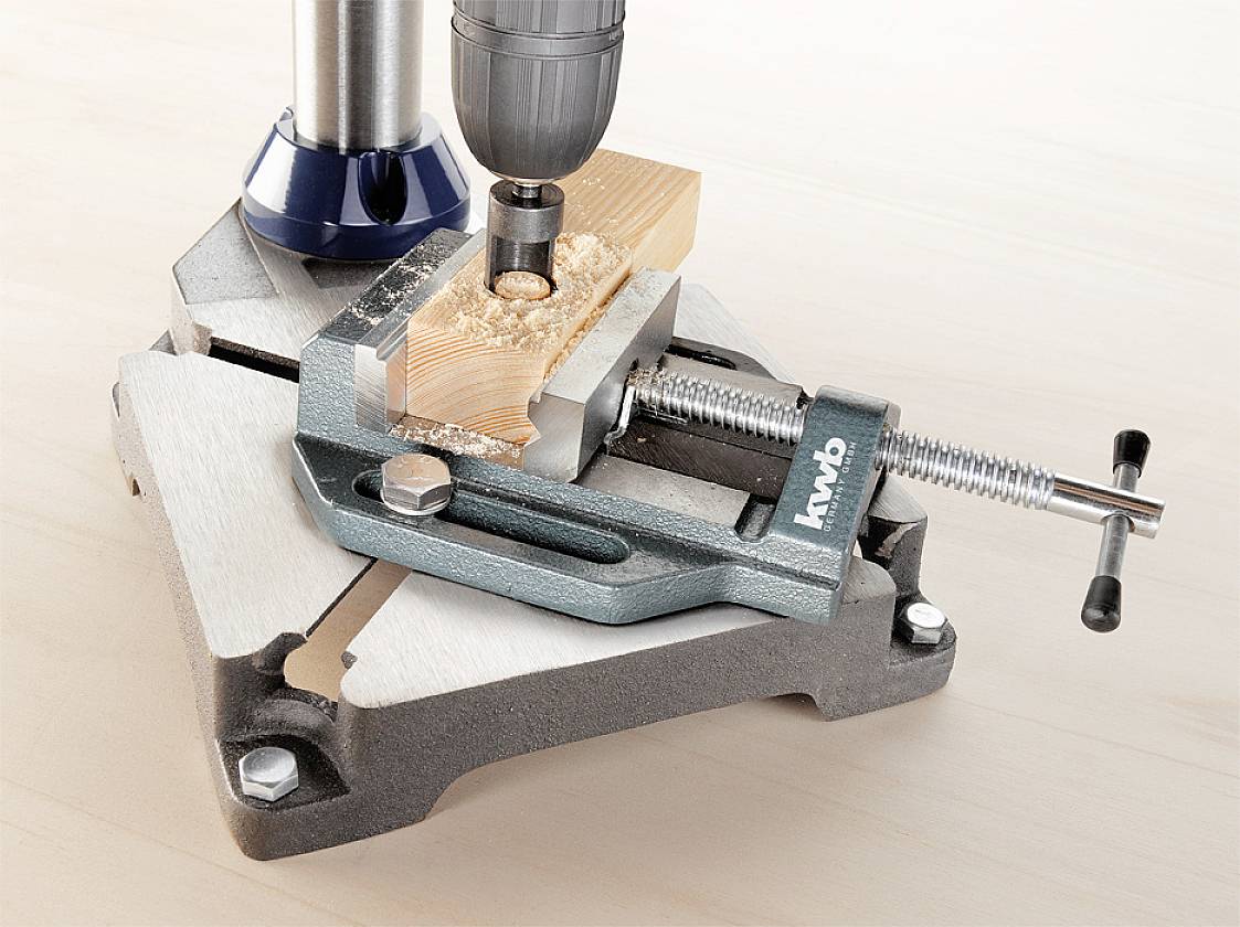 'Nahaufnahme einer Bohrmaschine, die ein Loch in ein Holzstück bohrt, das in einem Metall-Schraubstock auf einem Tisch fixiert ist.'