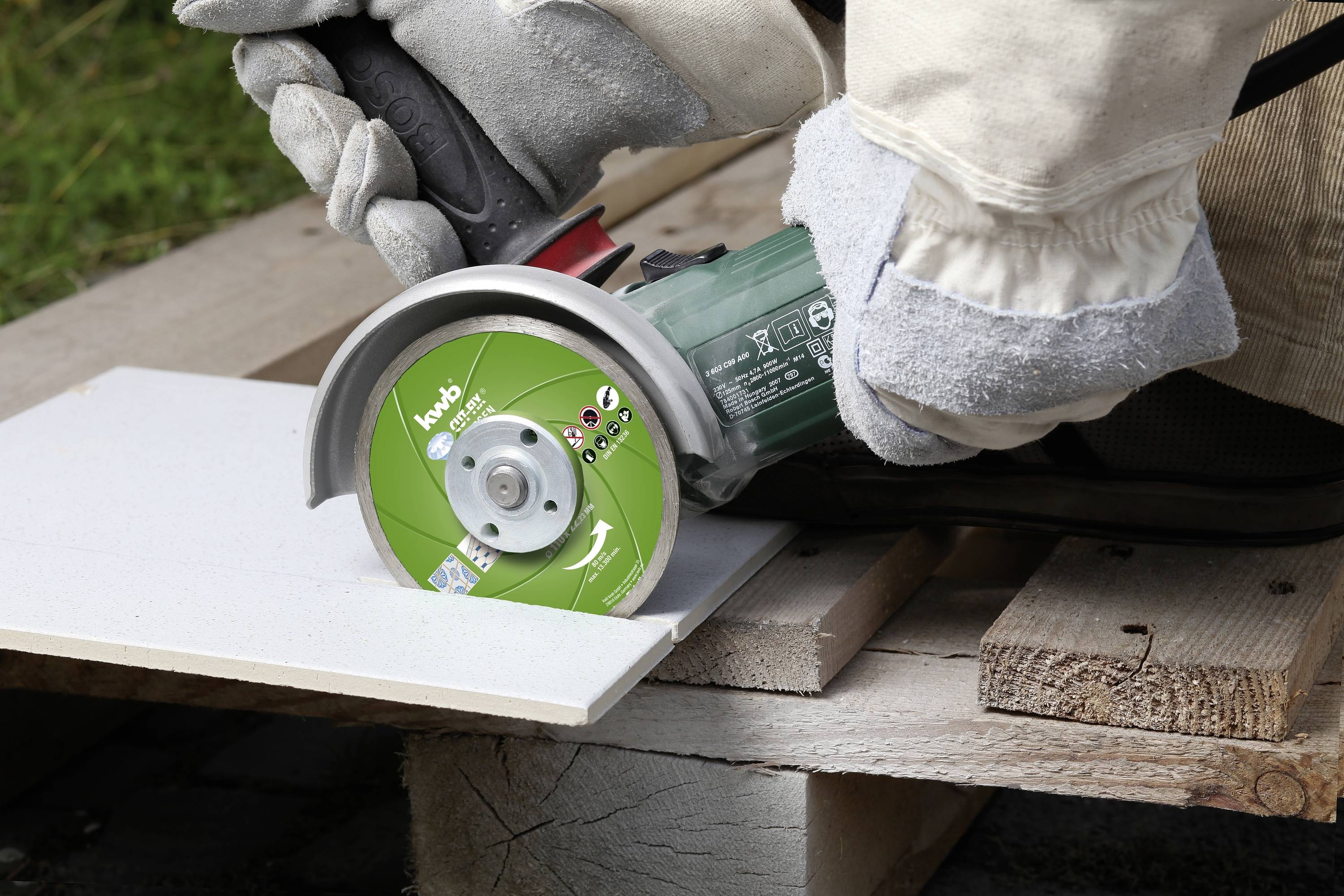 Eine Person schneidet mit einer Winkelschleifmaschine eine weiße Fliese. Sie trägt Sicherheitshandschuhe und schneidet auf einer Holzunterlage.