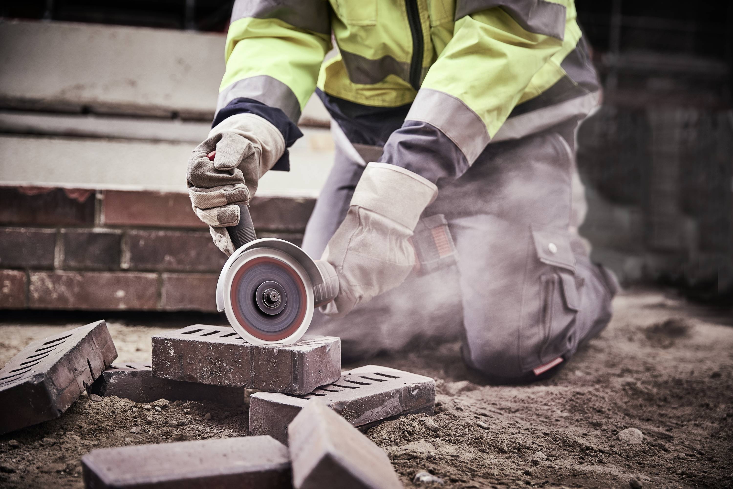 Eine Person in Sicherheitskleidung schneidet mit einer Winkelschleifer Steine. Staub steigt auf. Arbeitsplatz auf einer Baustelle.