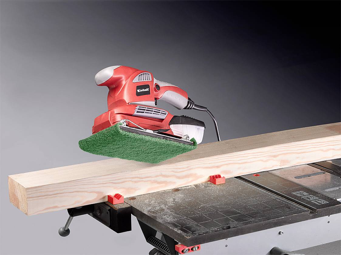 Ein roter Schwingschleifer schleift ein Holzbrett auf einem Tisch. Holzspäne sind sichtbar, das Werkzeug ist elektrisch.