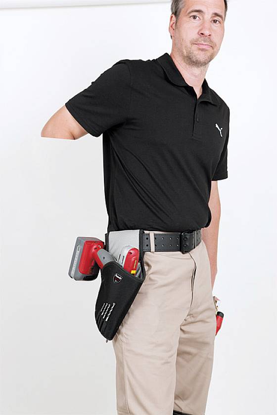 Ein Mann steht vor einem weißen Hintergrund und trägt ein schwarzes Poloshirt und beige Hosen. An seinem Gürtel hängt ein Werkzeughalter.