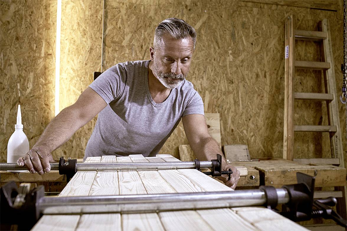 Ein Mann mittleren Alters arbeitet konzentriert an einem Holztisch in einer Werkstatt, umgeben von Werkzeugen und Holzmaterialien.