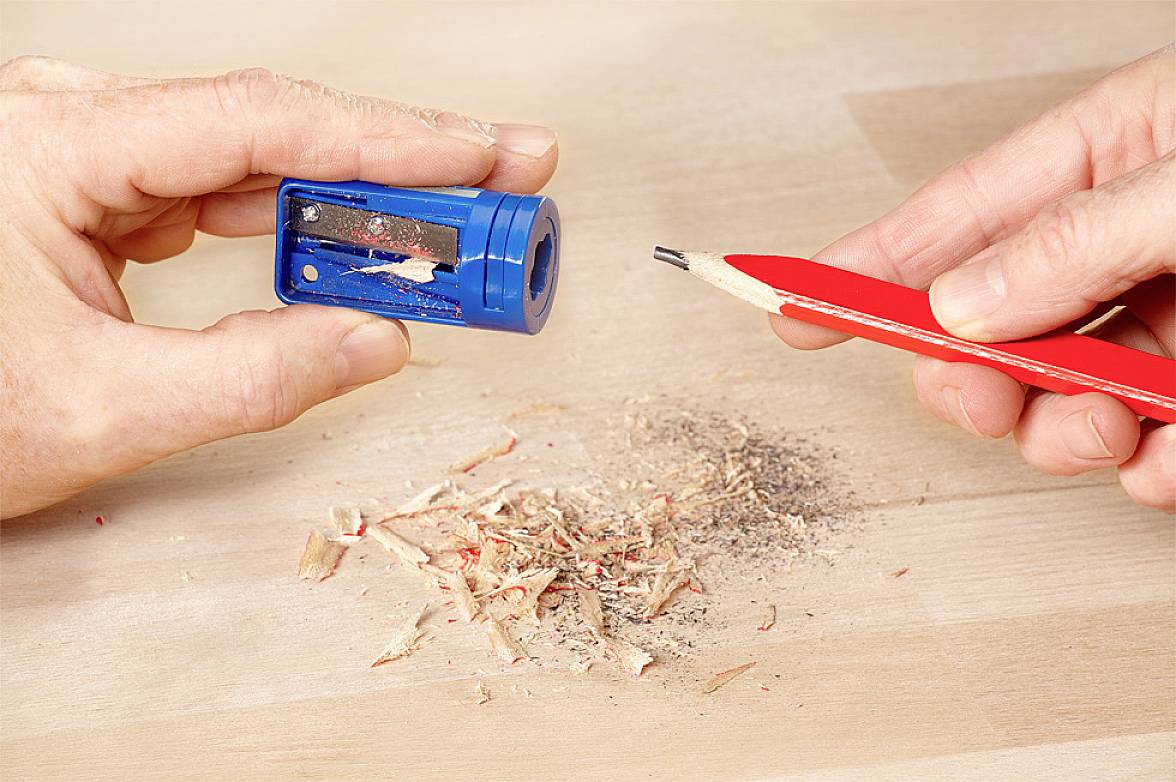 Eine Hand hält einen blauen Anspitzer, eine andere Hand einen roten Bleistift. Auf dem Tisch liegen Holzspäne vom Anspitzen.