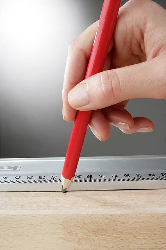 Ein roter Stift wird von einer Hand gehalten und zieht eine Linie entlang eines Lineals auf einem Holzbrett.