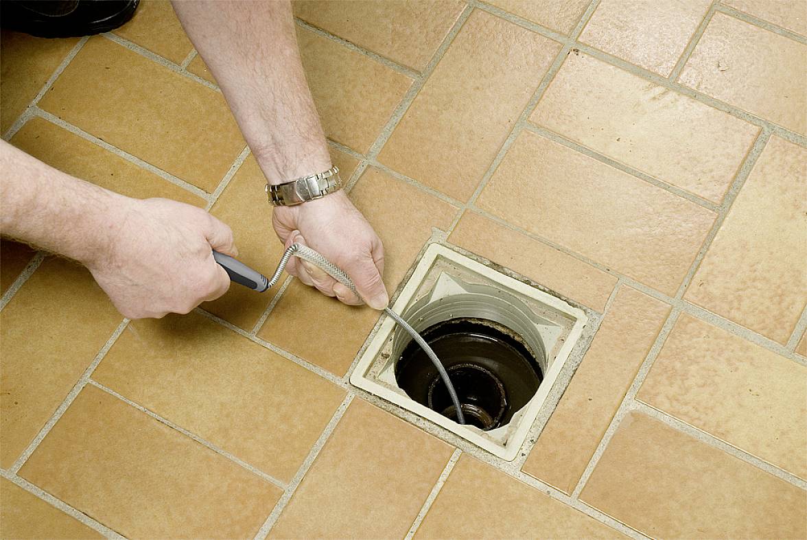 Eine Person reinigt einen Abfluss auf einem gefliesten Boden mit einem flexiblen Kabelwerkzeug, um Verstopfungen zu entfernen.