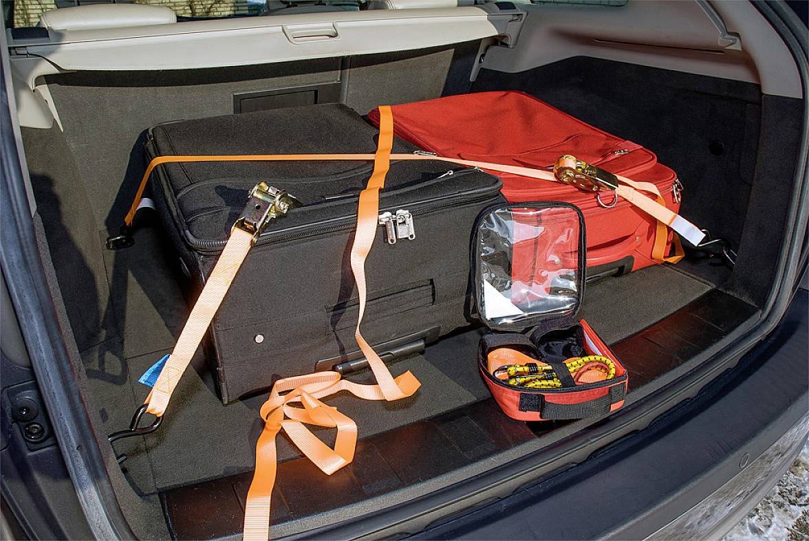Gepäck gesichert im Kofferraum eines Autos: Zwei Koffer mit Spanngurten befestigt, ein offener Werkzeugkasten zeigt Spannmeter und Gurte.