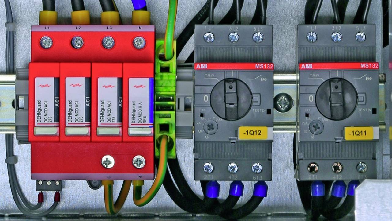 Ein elektrischer Schaltkasten mit Schaltgeräten und Sicherungen, darunter ein roter mehrpoliger Hauptschalter und zwei graue Schutzschalter.