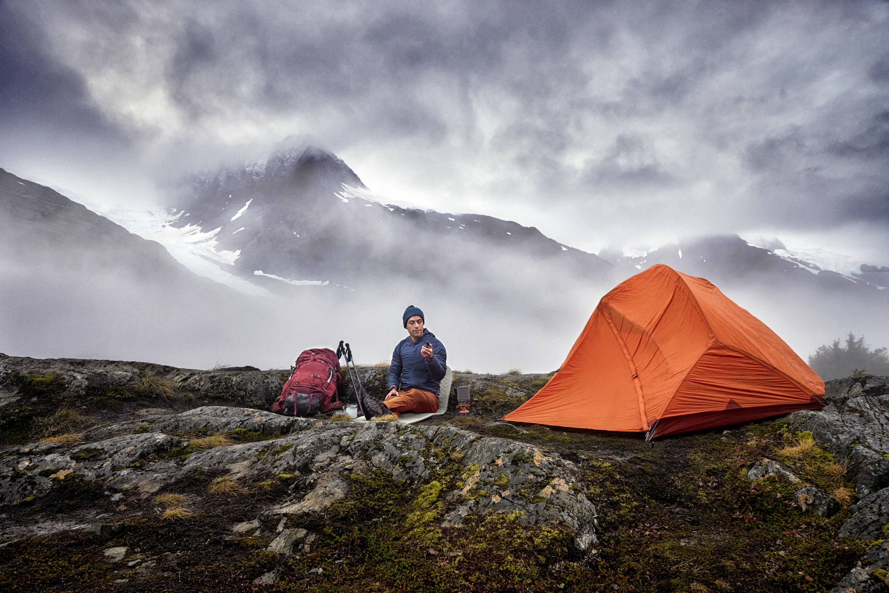 Eine Person sitzt neben einem orangefarbenen Zelt in einer bergigen, nebligen Landschaft. Ein Rucksack steht daneben.