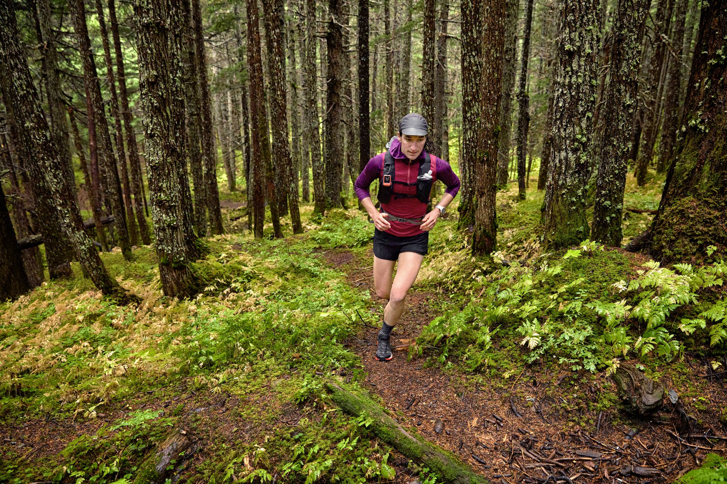 Eine Person läuft durch einen dichten Wald auf einem schmalen, mit Moos bedeckten Pfad, umgeben von hohen Bäumen und üppigem Grün.