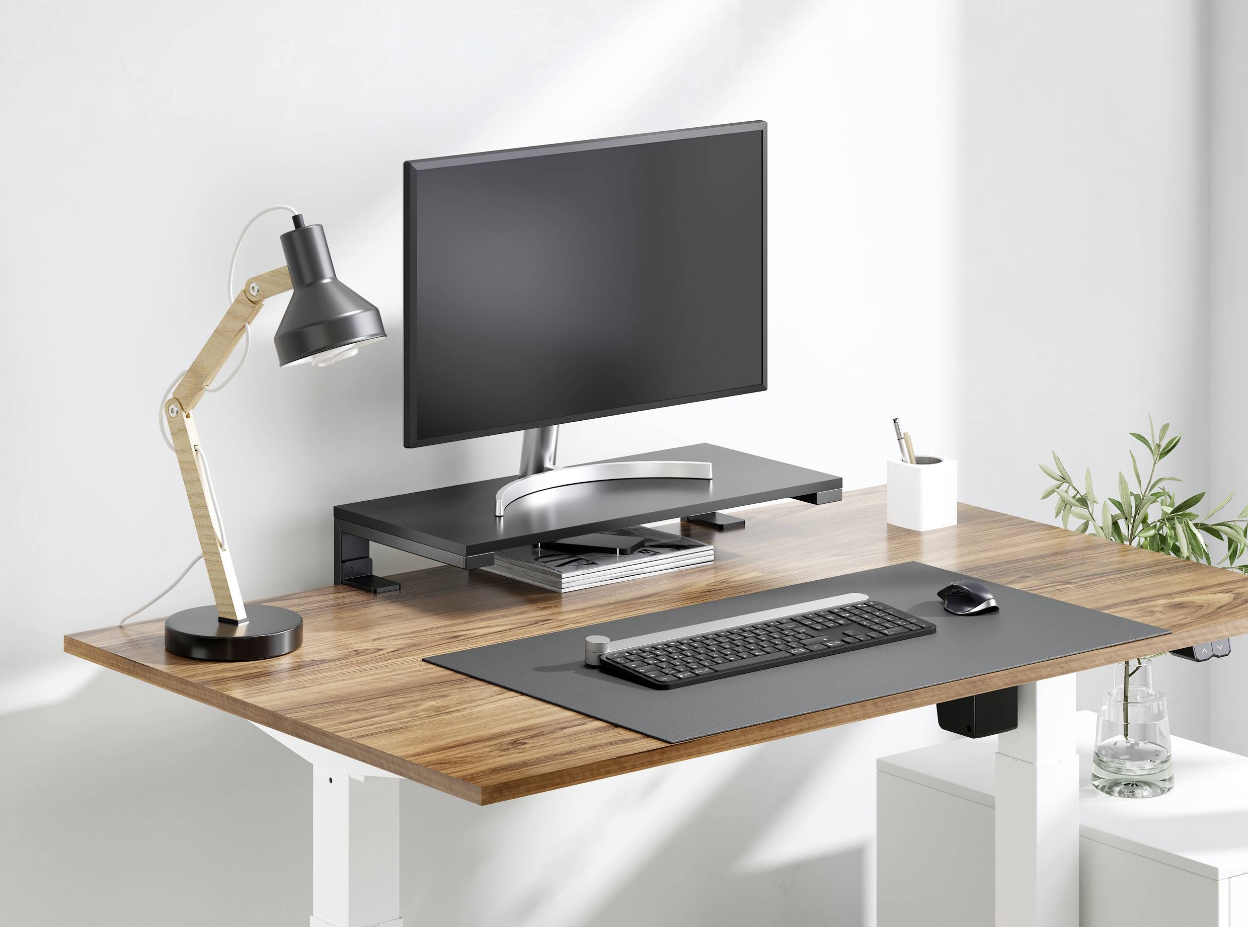 Moderner Schreibtisch mit Holzoberfläche, Monitor, Tastatur, Maus und Schreibtischlampe. Im Hintergrund sind Pflanzen zu sehen.