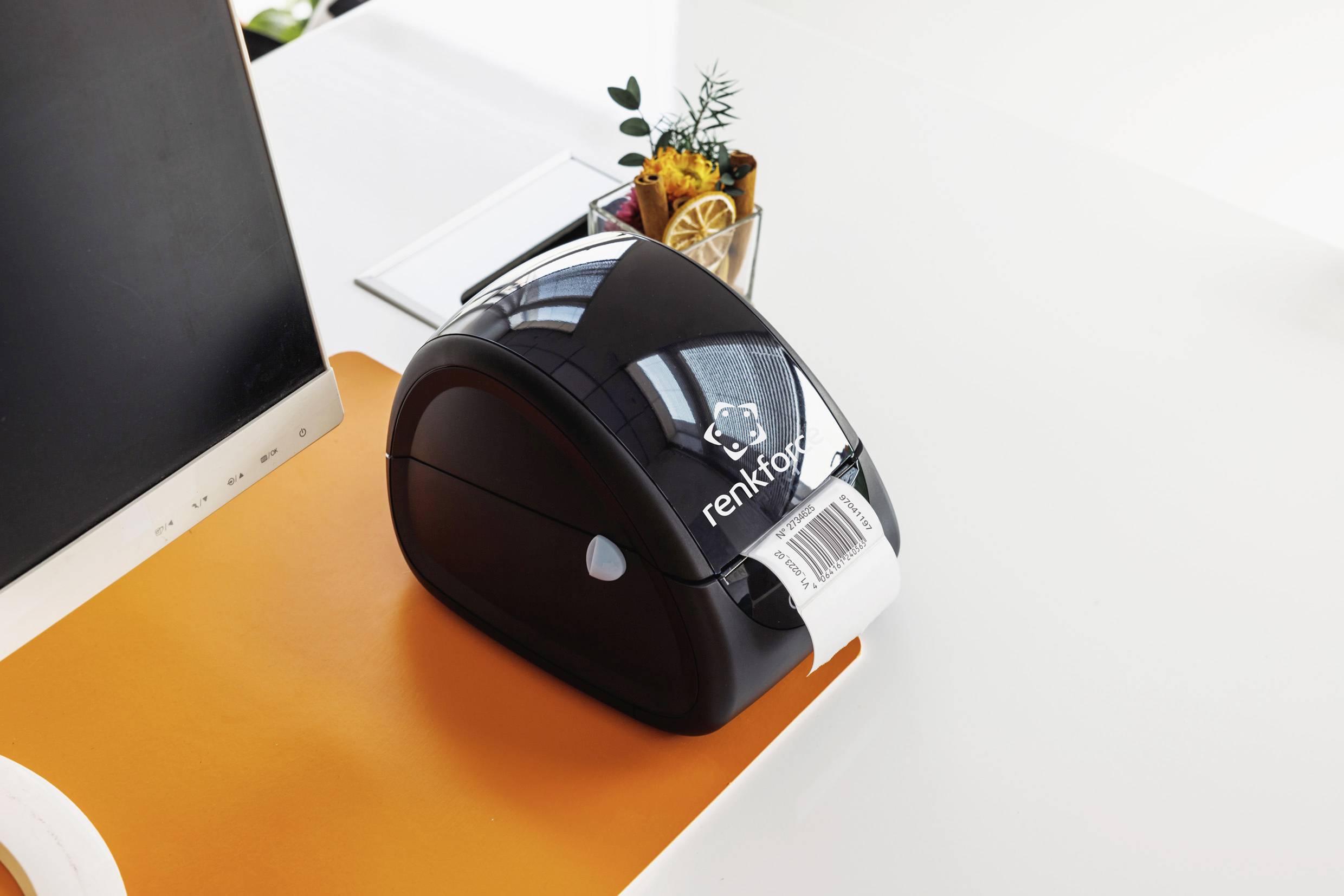 Ein kleiner schwarzer Drucker sitzt auf einem orangefarbenen Schreibtisch und druckt ein weißes Etikett. Im Hintergrund sind Pflanzen.