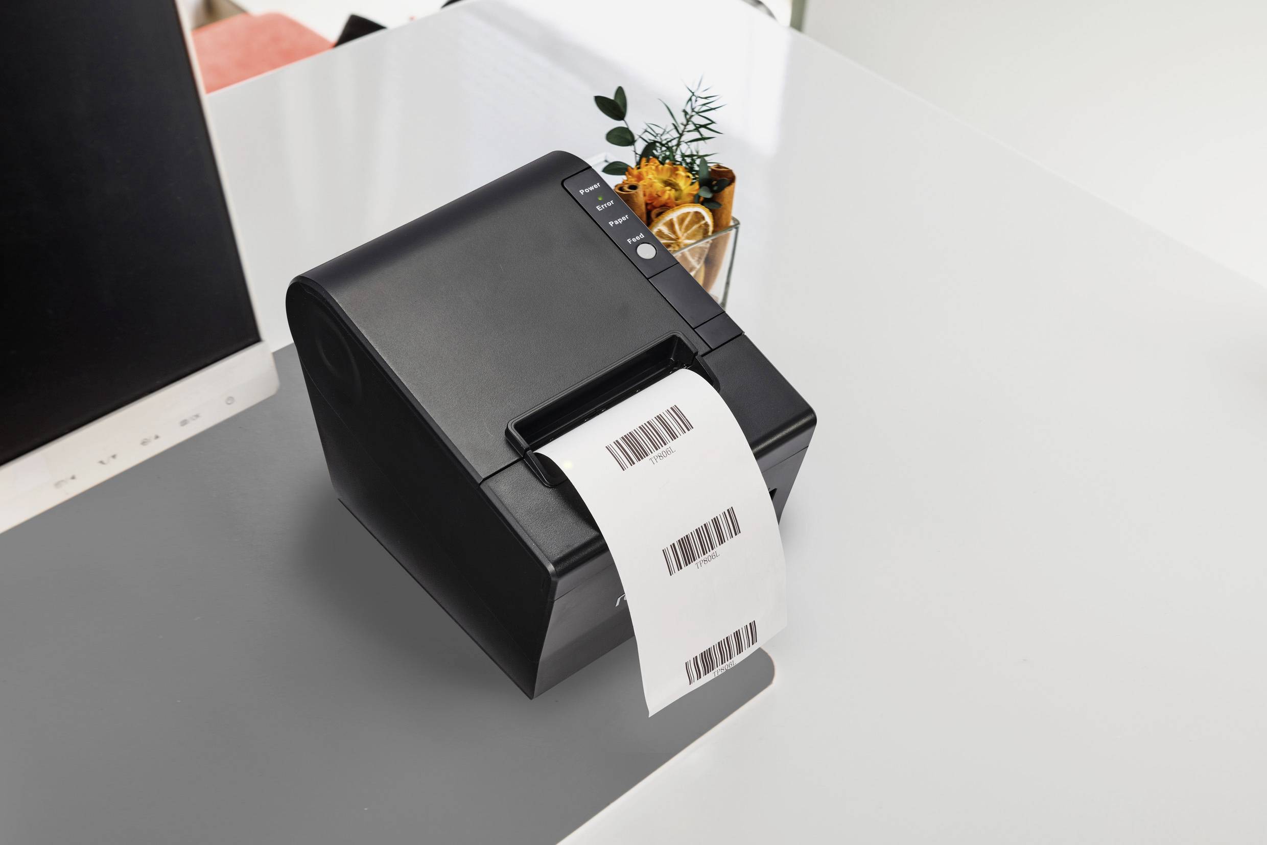 Ein schwarzer Etikettendrucker auf einem weißen Tisch druckt ein Blatt mit Barcodes. Im Hintergrund ist ein kleiner Blumentopf sichtbar.