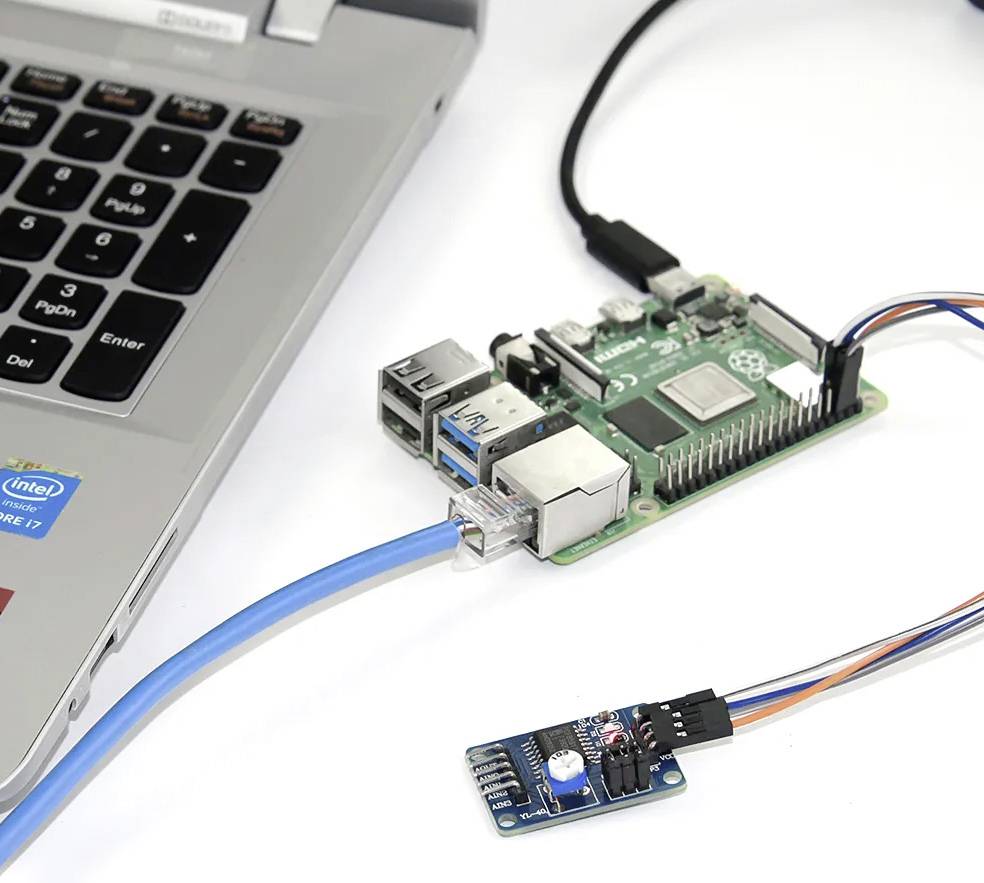 Ein Raspberry Pi, über ein Netzwerkkabel mit einem Laptop verbunden, neben einem kleinen Sensor mit bunten Kabeln auf weißem Hintergrund.