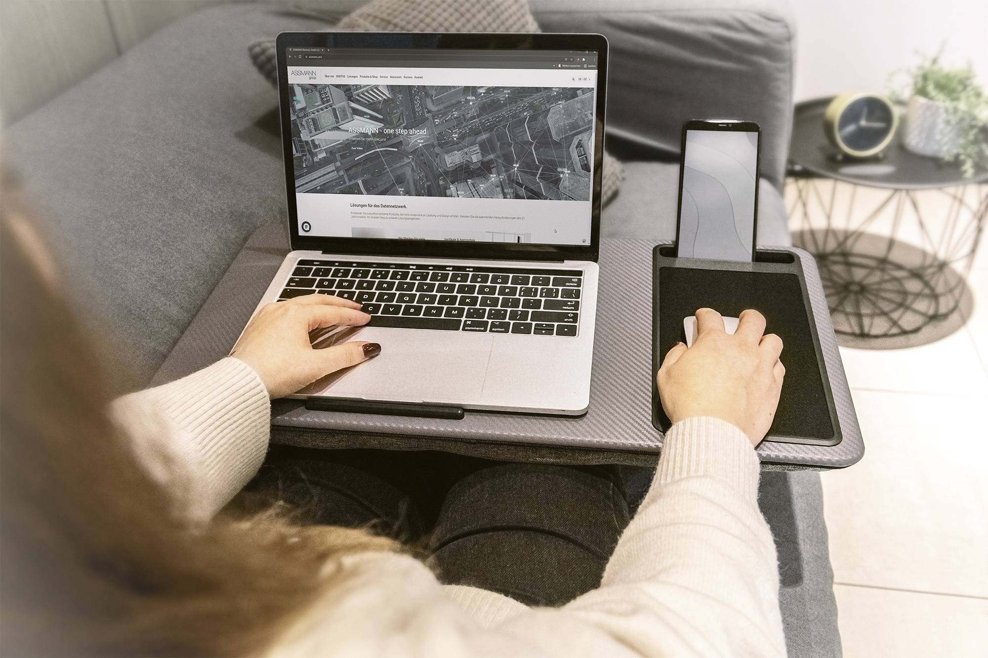 Eine Person arbeitet auf einem Laptop mit einer Webseite geöffnet, daneben ein Smartphone auf einem Sofa. Sie verwendet eine externe Maus.