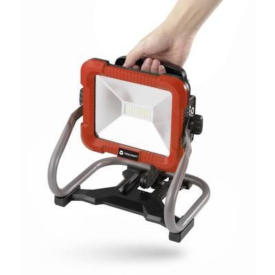 Eine Hand hält einen tragbaren, rot-schwarzen LED-Baustrahler mit einem stabilen Ständer, geeignet für den Einsatz auf Baustellen.