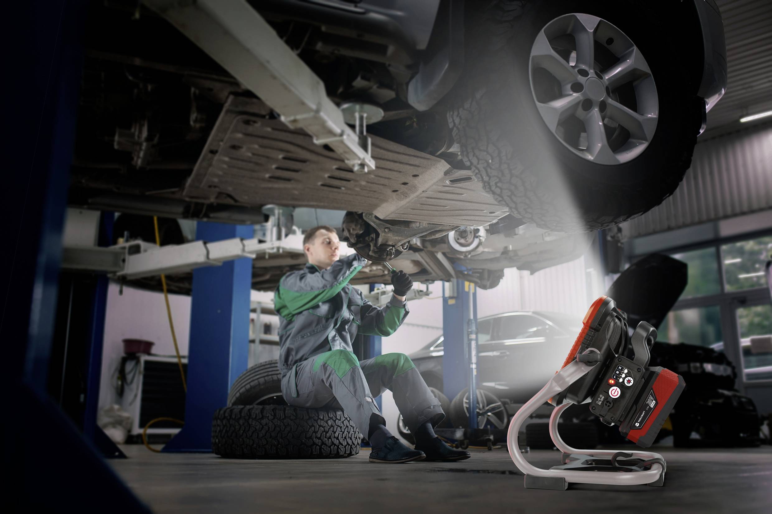 Ein Mechaniker repariert unter einem aufgebockten Auto in einer Werkstatt. Eine Lampe erhellt den Arbeitsbereich. Reifen sind sichtbar.