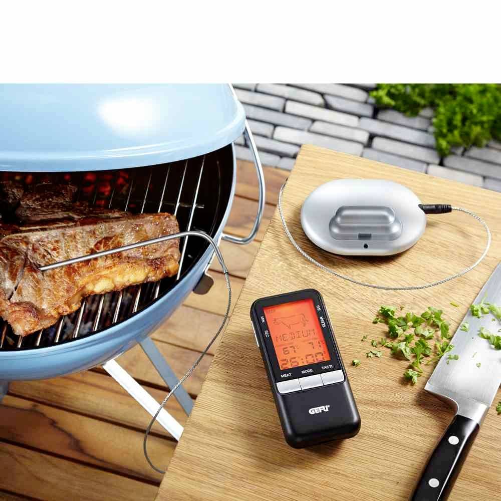 Grill mit Fleisch and angeschlossenem Fleischthermometer, das die Temperatur anzeigt; daneben Kräuter und Messer auf Holzbrett.