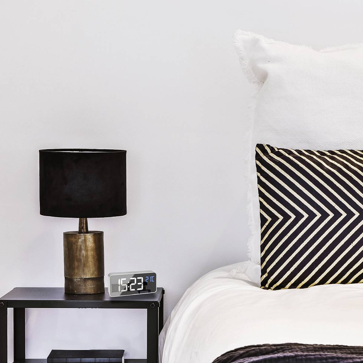 Ein Schlafzimmer mit modernem Nachttisch, schwarzer Lampe, Digitaluhr (23:23 Uhr), beigem Kissen mit Zickzack-Muster, weißem Kissen und Bettdecke.
