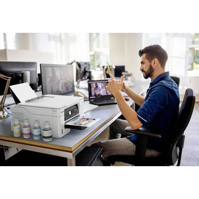 Ein Mann sitzt in einem Büro an einem Schreibtisch, bedient einen Laptop und schaut auf den Bildschirm. Neben ihm steht ein Drucker.