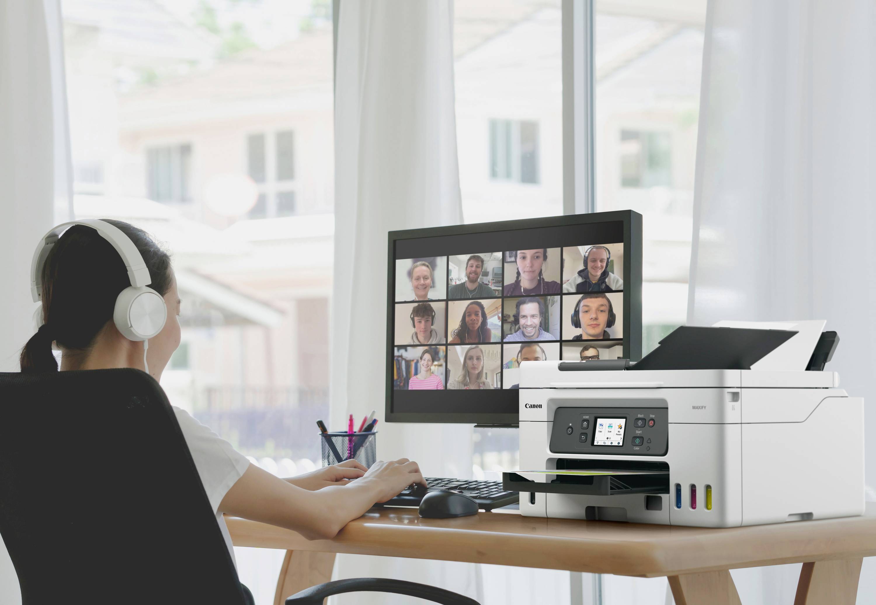 Eine Person sitzt am Schreibtisch mit Kopfhörern und nimmt an einem Videomeeting auf einem Computerbildschirm teil. Rechts daneben steht ein Drucker.