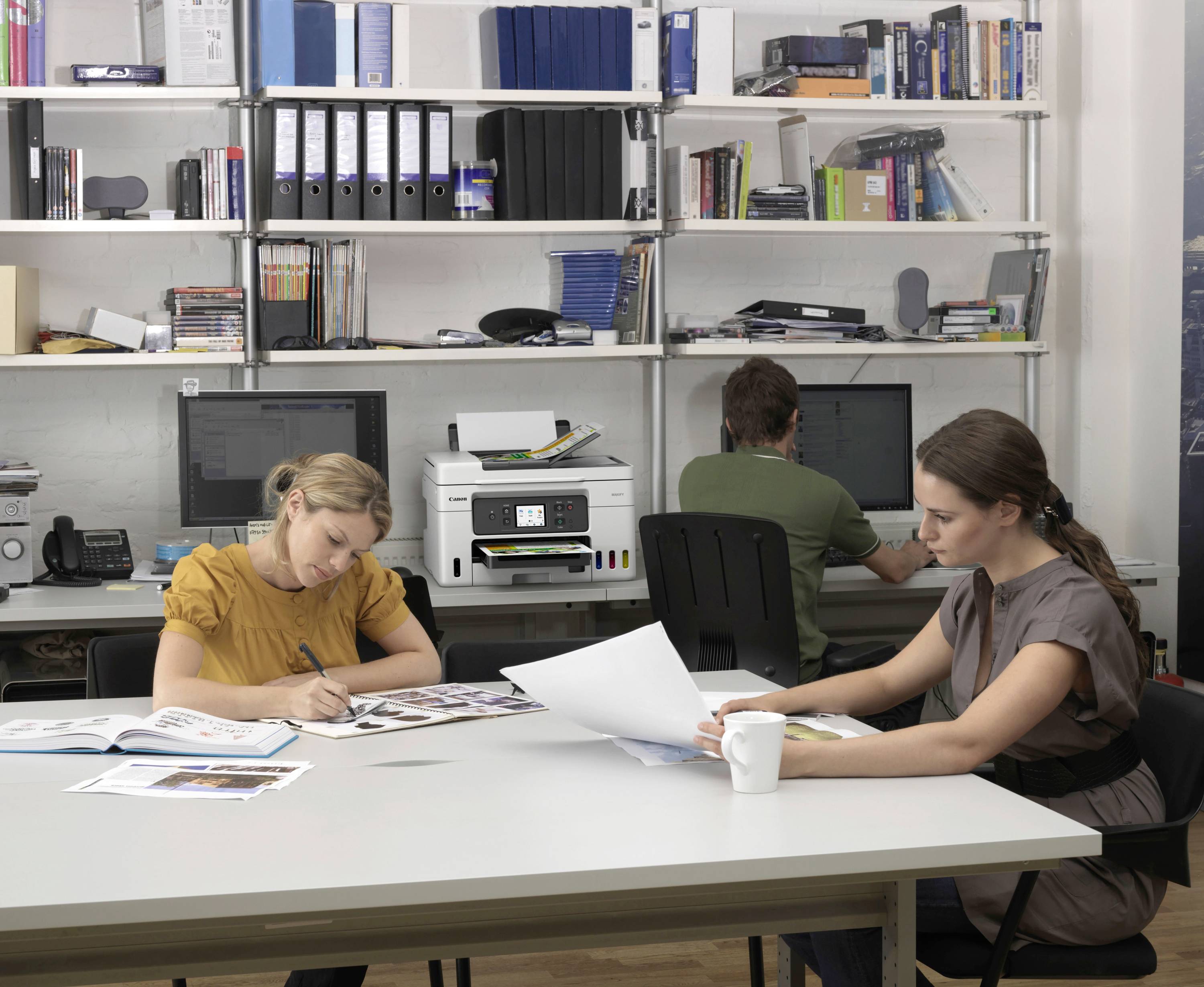 Drei Personen arbeiten an einem Tisch in einem Büro. Zwei Frauen studieren Dokumente, während ein Mann an einem Computer sitzt. Regale voller Aktenordner im Hintergrund.