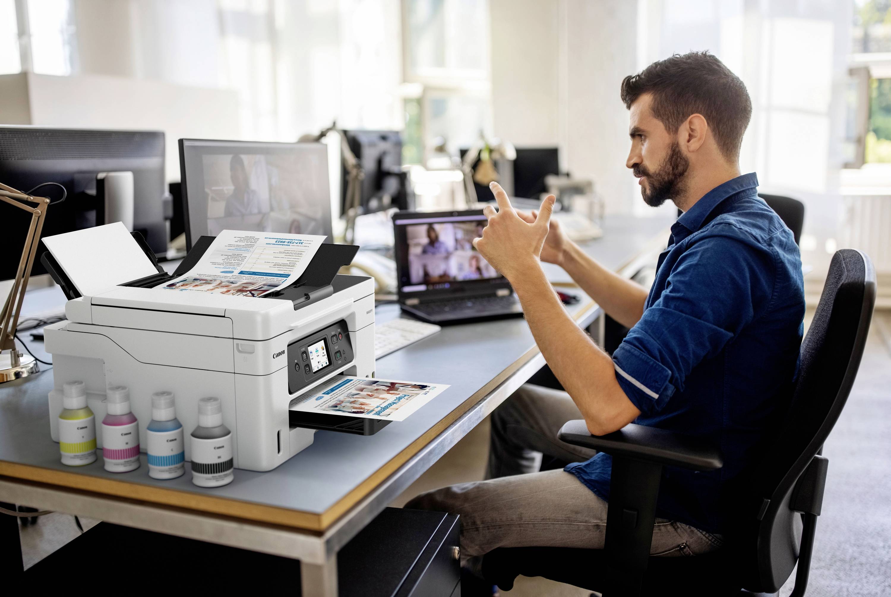Eine Person sitzt an einem Schreibtisch und gebärdet vor einem Laptop. Ein Drucker druckt Dokumente. Im Hintergrund sieht man ein Büro.