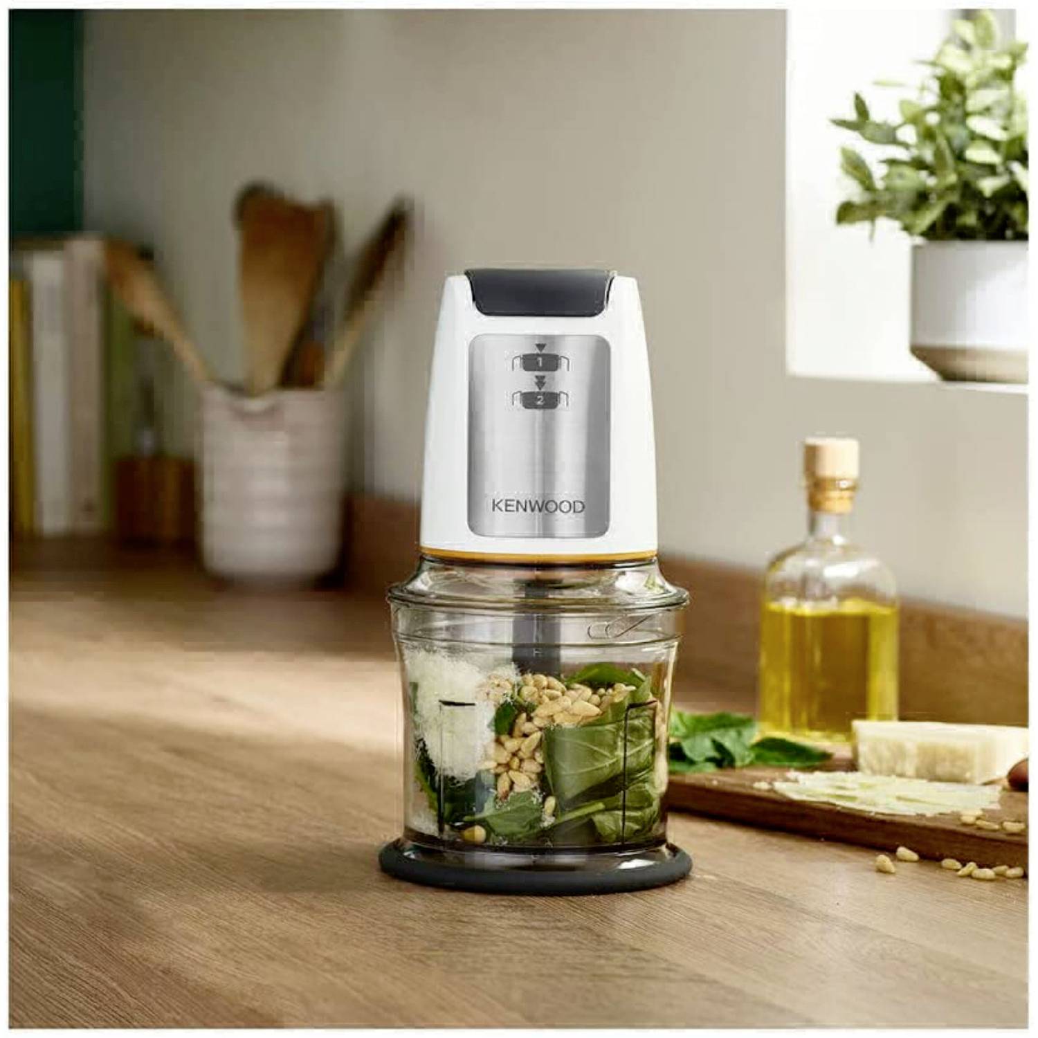 Ein Küchenmixer auf einer Arbeitsplatte enthält Gemüse und Nüsse. Im Hintergrund sind Küchenutensilien und Gewürze sichtbar.