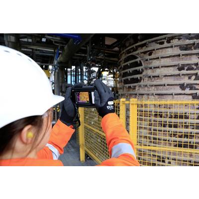 Eine Person in Schutzkleidung fotografiert eine industrielle Anlage mit einer Wärmebildkamera. Eine große Metallstruktur ist sichtbar.