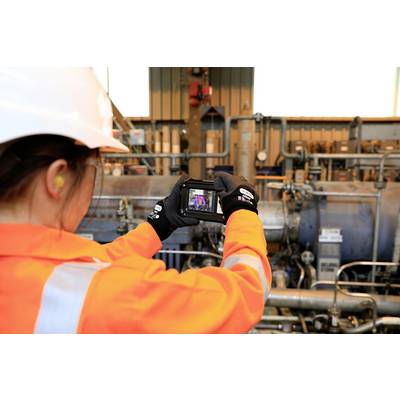 Eine Person in orangefarbener Schutzkleidung und Helm verwendet ein Kameragerät in einer industriellen Umgebung mit Rohren und Maschinen.