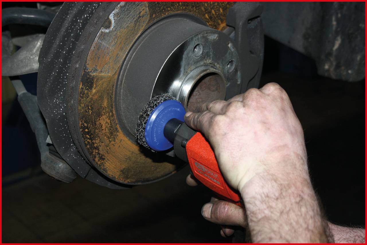 Eine Person reinigt eine rostige Bremsscheibe mit einem roten Elektrowerkzeug.
