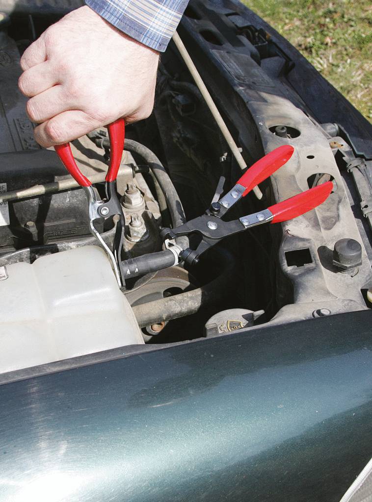 Eine Person hält eine Zange im Motorraum eines Autos, um an einem Teil zu arbeiten. Hände und Werkzeug im Fokus.