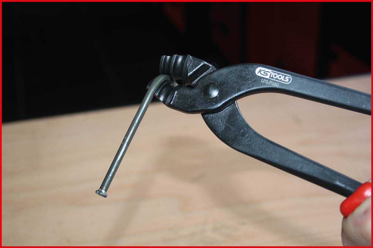 Eine schwarzmetallische Zange biegt einen langen Nagel auf einem hellen Holzuntergrund. Das Werkzeug hat die Aufschrift 'GS TOOLS'.