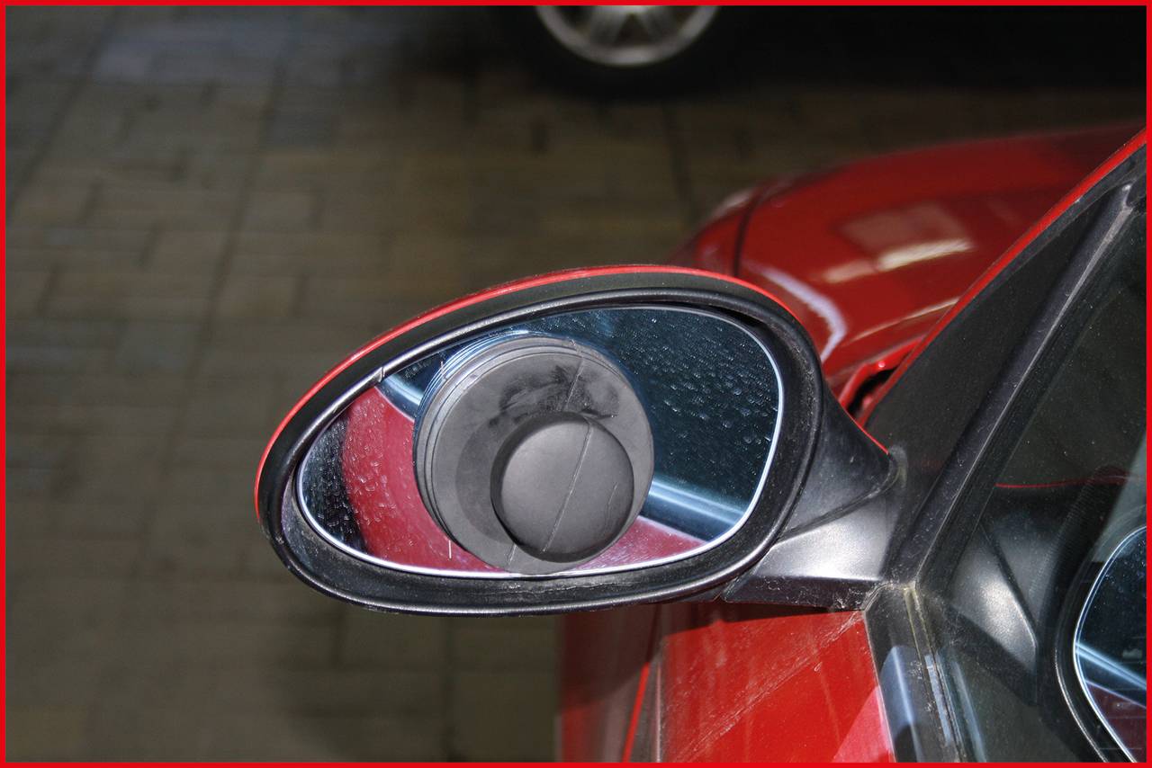 Seitenspiegel eines roten Autos mit Kratzern und Spuren.
