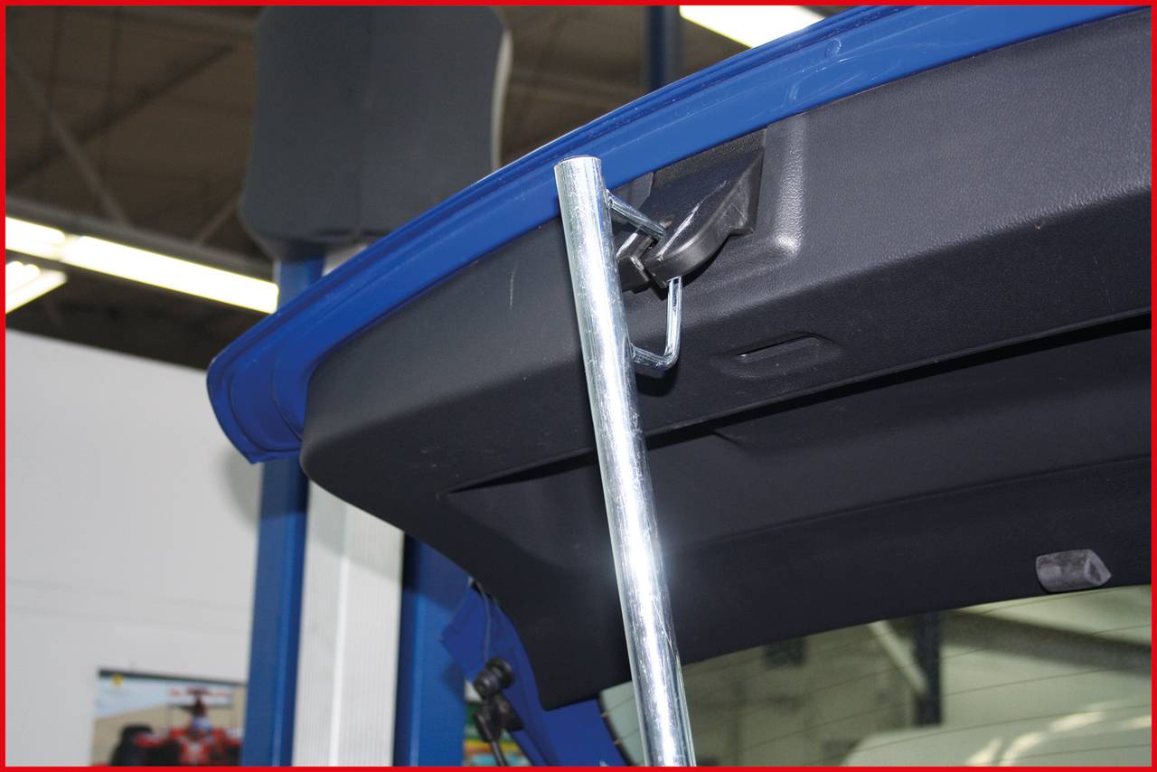 Ein blauer Kofferraumdeckel, gestützt von einer Metallstange in einer Werkstatt. Im Hintergrund sind unscharfe Werkstattelemente erkennbar.