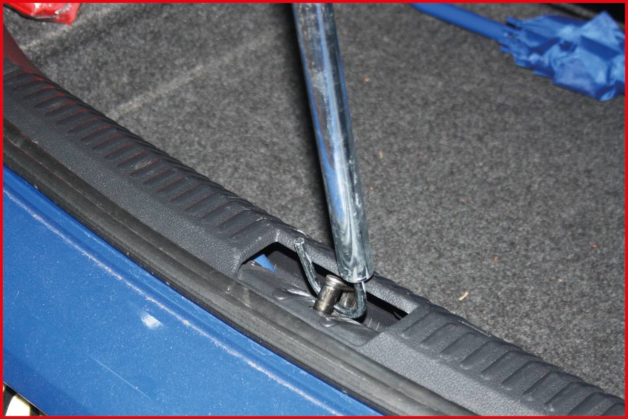 Ein Wagenheber ist an einem Auto im Kofferraum installiert. Ein blauer Regenschirm ist im Hintergrund zu sehen.